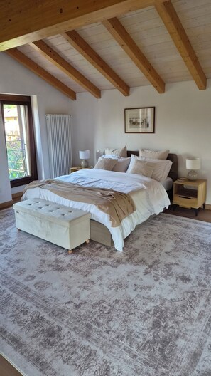 A bedroom with a large bed, a bedside table, a nightstand, a radiator, a window, and a wooden ceiling.