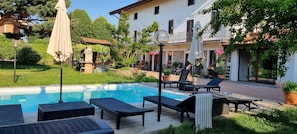 A pool area with lounge chairs and umbrellas, surrounded by greenery and a building with stone walls.