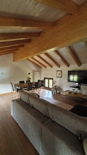 Ein geräumiges Wohnzimmer mit hohen Decken, Holzbalken, einem großen Sofa, einem Esstisch, Stühlen, einem Fernseher und einer Steinskulptur.