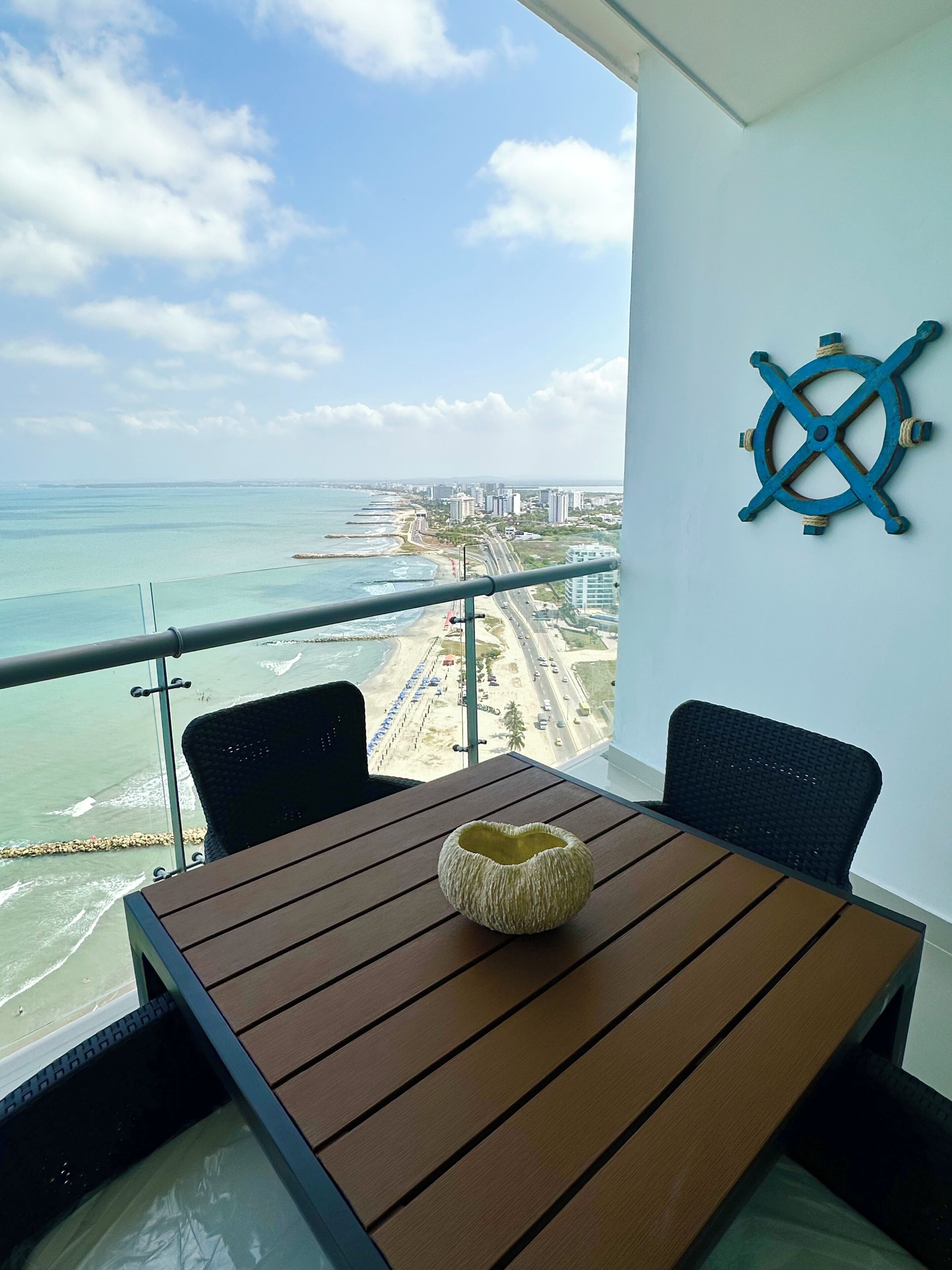 A balcony with a view of the beach and ocean, a table with chairs, and a ship's wheel decoration.