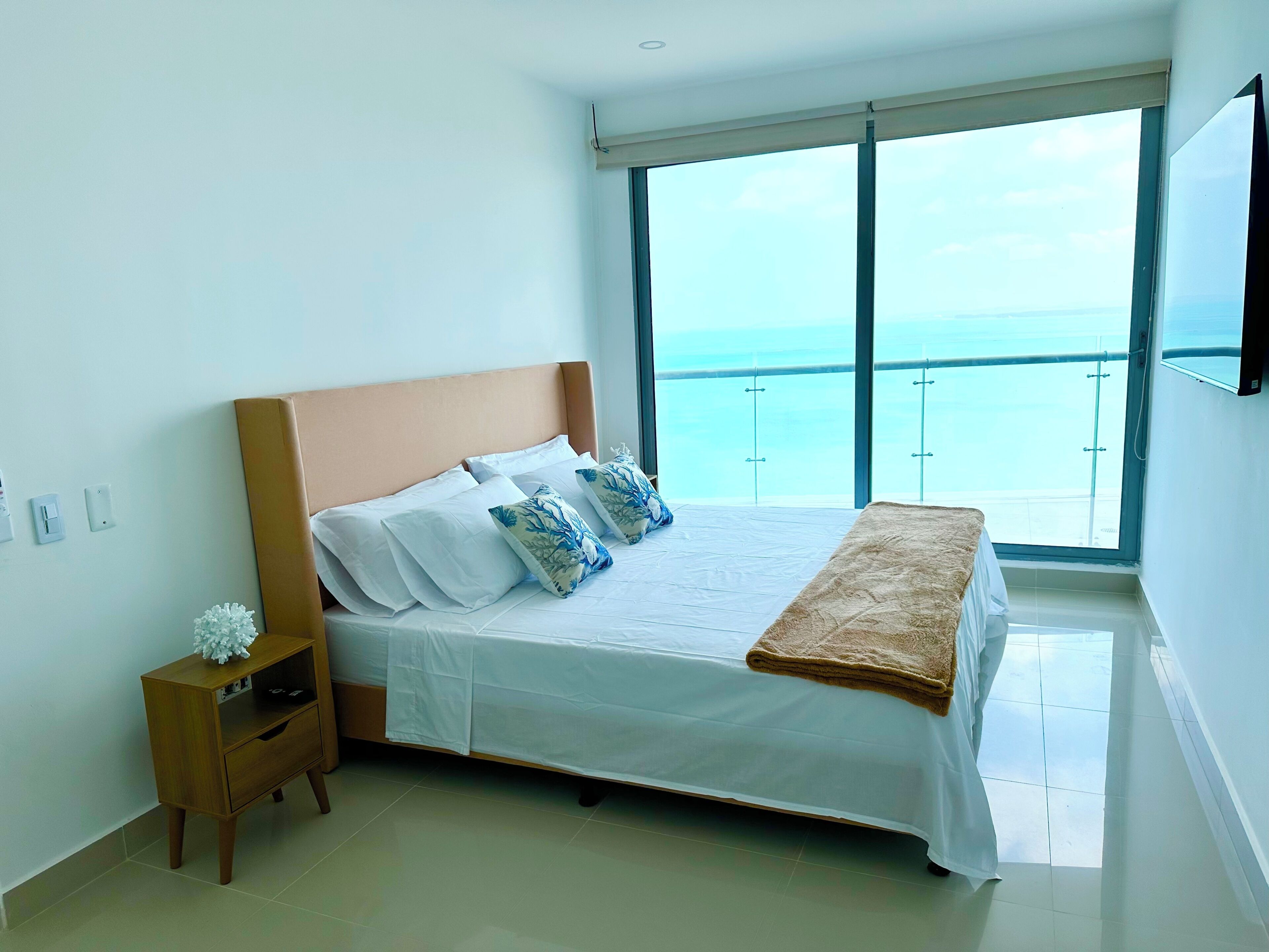 A bedroom with a large bed, a nightstand, a TV, and a view of the sea through a balcony.