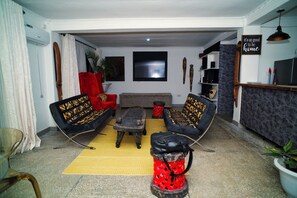 Un salon comprenant un canapé, une table basse et un tabouret rouge. Un téléviseur à écran plat est fixé au mur.