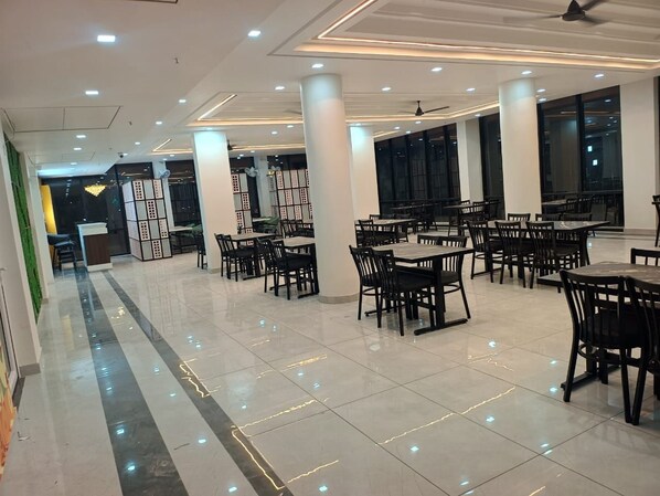 A spacious indoor area with multiple dining tables and chairs, large windows, and a modern ceiling design.