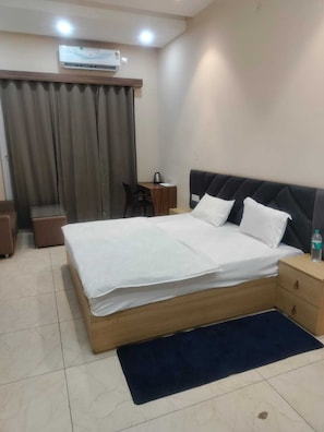 A hotel room with a bed, bedside table, chair, and an air conditioning unit. - Hotel Kanti Empire (Dholpur)
