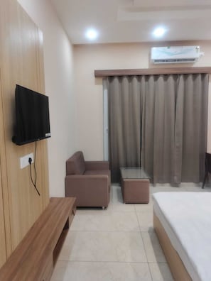 A hotel room with a bed, a sofa, a TV mounted on the wall, an air conditioning unit, and a window with curtains. - Hotel Kanti Empire (Dholpur)