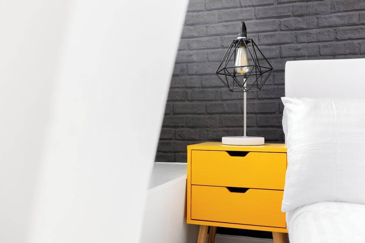 En seng med hvide sengetøj, et gult natbord med skuffer og en sort geometrisk lampe på bordet.