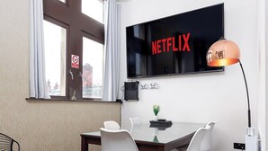 Ett modernt rum med en stor TV som hänger på väggen och visar "NETFLIX", ett matbord med stolar, en liten växt och en spelkonsol.