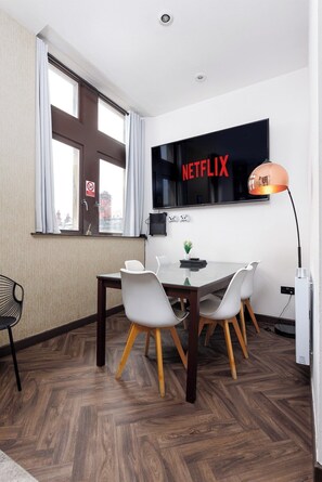 A modern room with a large wall-mounted TV displaying 'NETFLIX,' a dining table with chairs, a small plant, and a gaming console. - Flatzy - Stunning Victorian Flat in Centre + Xbox (Liverpool)