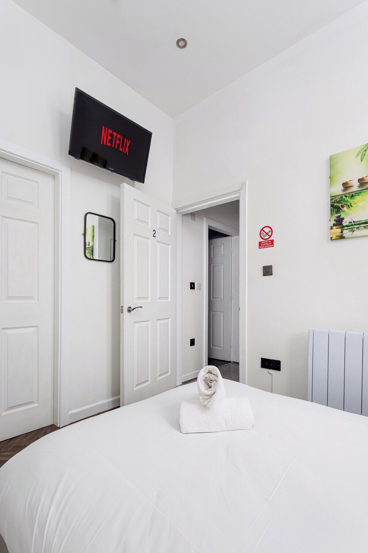 Habitación de hotel con una cama, un televisor montado en la pared, una puerta con un número y un cuadro en la pared.