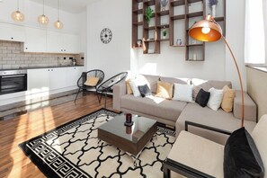 A modern living room with a sofa, coffee table, and chairs, a kitchen in the background, and a large bookshelf. - Flatzy - Stunning Victorian Flat in Centre + Xbox (Liverpool)