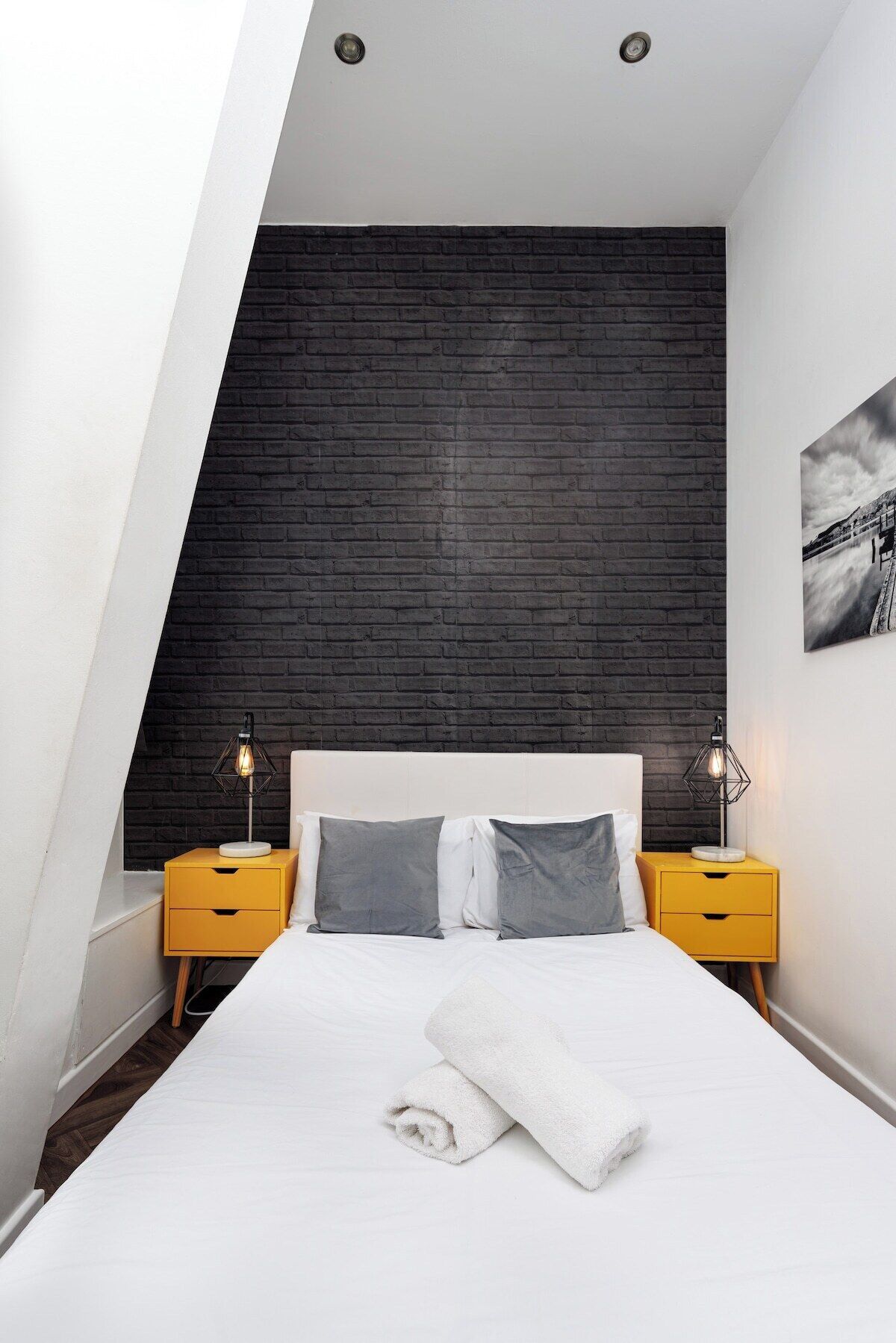 A modern bedroom with a sloped ceiling, a white bed, two nightstands, and a black brick accent wall.