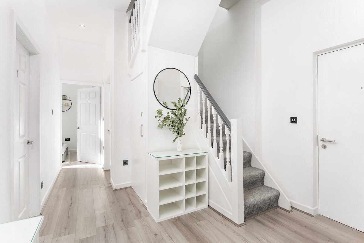 A hallway with a staircase, a white wall with a mirror, and a plant on a glass shelf.