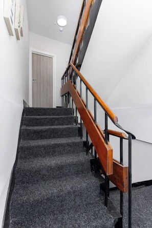 A staircase with a wooden handrail and black metal balusters, leading to a door with a light switch above it. - Flatzy - 16 Guests 4 En-suite Central Apartment B (Liverpool)