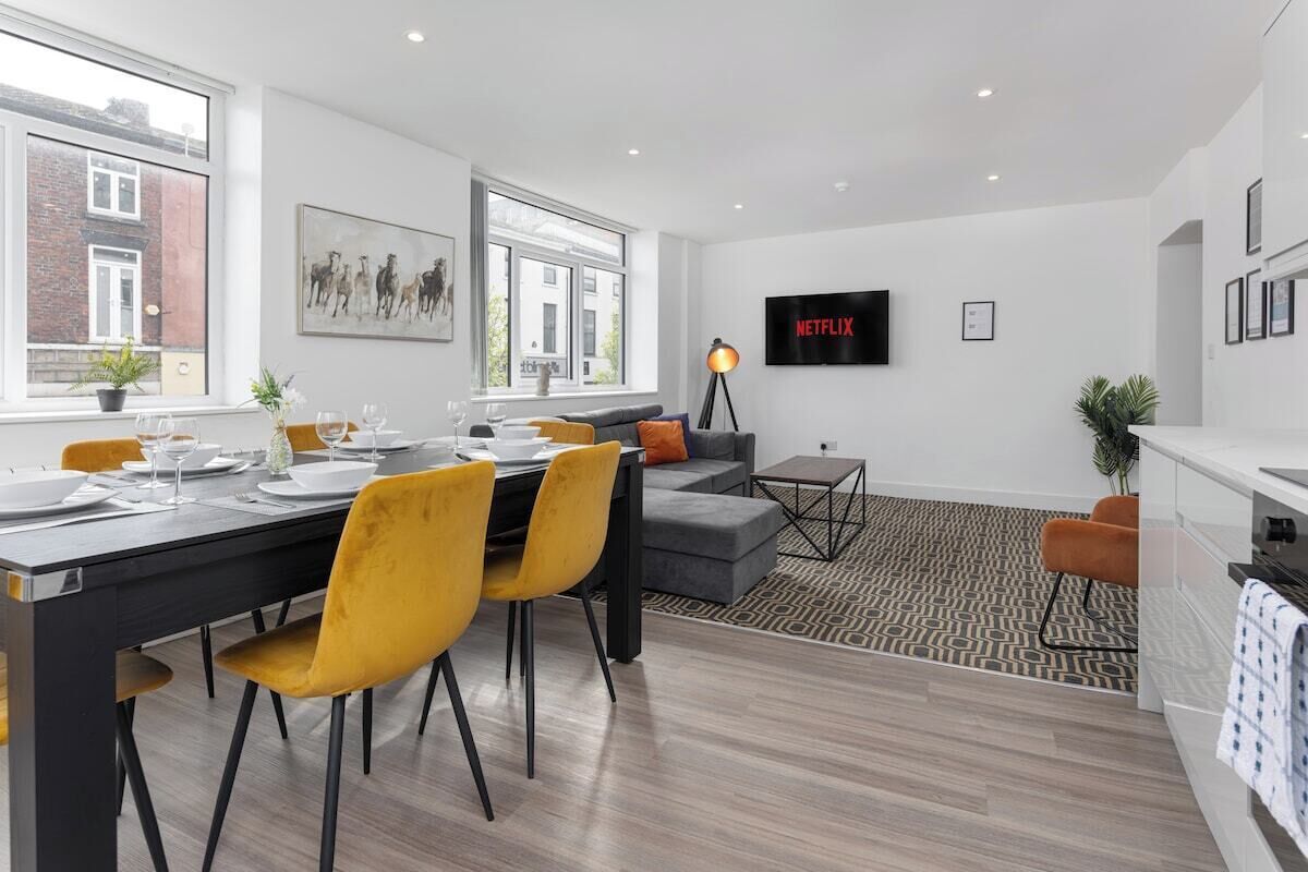 A modern living room with a dining area, a grey sofa, a TV mounted on the wall, and a patterned rug.