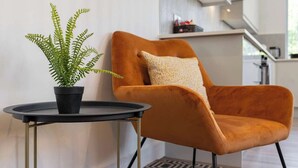 Ein modernes Wohnzimmer mit einem einzelnen orangefarbenen Sessel, einem kleinen runden Tisch und einem Topf mit einem Farne.