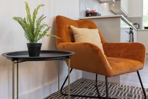 A modern living room with a single orange armchair, a small round table, and a potted fern. - Flatzy - 16 Guests 4 En-suite Central Apartment B (Liverpool)