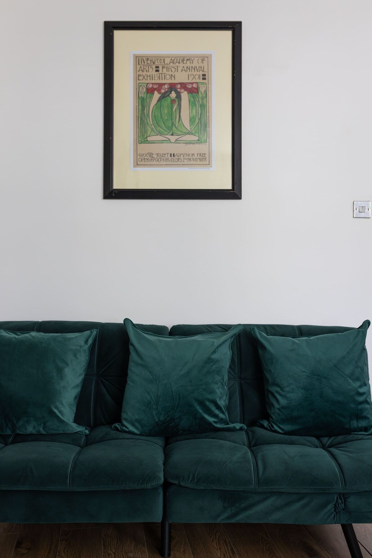 A framed artwork on a wall above a dark green sofa.