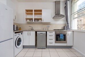 A modern kitchen with a washing machine, dishwasher, oven, and a window with a view of buildings. - Flatzy - City Centre  Modern 2 bedroom Apartment (Merseyside)