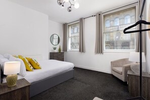A modern hotel room with a bed, bedside table, lamp, chair, and large windows with curtains. - Flatzy - City Centre  Modern 2 bedroom Apartment (Merseyside)