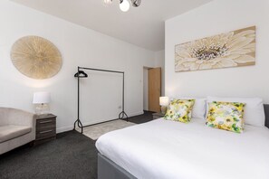 Ein Schlafzimmer mit Bett, Stuhl, Nachttisch, Lampe und Wandbild.