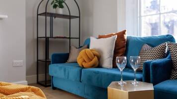 Ein Wohnzimmer mit einem blauen Sofa, einem gelben Hocker, einem schwarzen Metallregal mit einer Pflanze und zwei Weingläsern auf einer Holzbox.