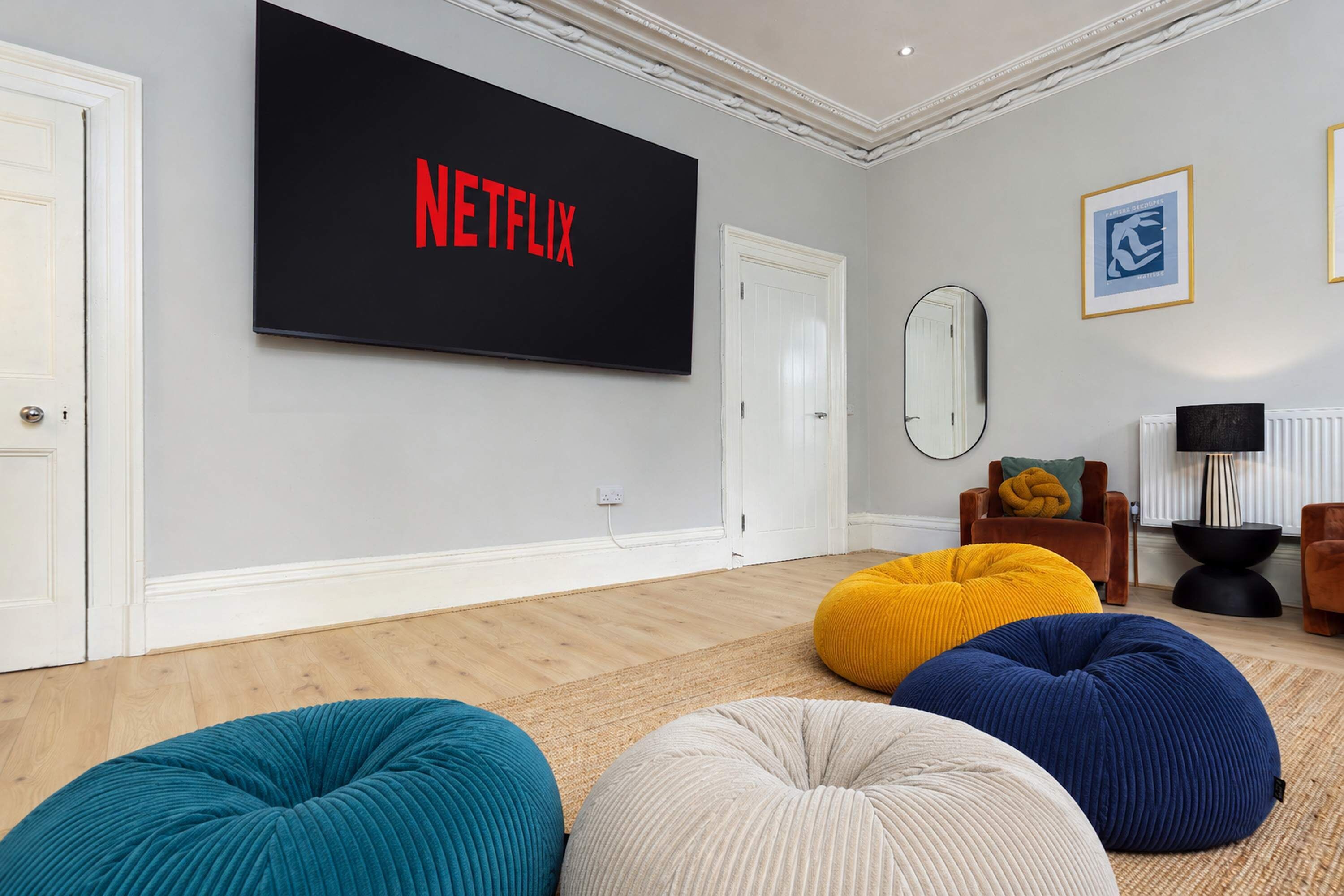 Ein Wohnzimmer mit einem großen Flachbildfernseher an der Wand, der 'NETFLIX' in roten Buchstaben anzeigt, einem Spiegel und einer Couch mit bunten Sitzsäcken.