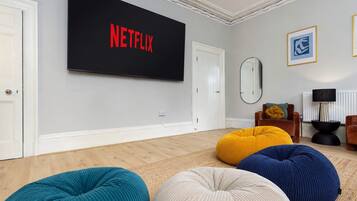 Ein Wohnzimmer mit einem großen Flachbildfernseher an der Wand, der 'NETFLIX' in roten Buchstaben anzeigt, einem Spiegel und einer Couch mit bunten Sitzsäcken.