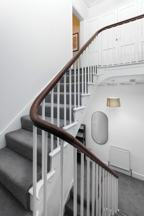 A staircase with white railings and dark wooden handrails, leading to a landing with a light fixture and a radiator. - Flatzy - City Centre Mansion | Sleeps 22 | 5 Baths (Liverpool)