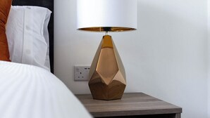 Ein Bett mit weißer Bettwäsche und einem braunen Kopfkissen, ein Nachttisch aus Holz mit einer geometrischen Lampe und ein gemustertes Teppich.