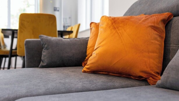 Ein graues Sofa mit zwei Kissen, ein orangefarbenes und ein graues, in einem Raum mit einem gelben Sessel und einem Fenster im Hintergrund.