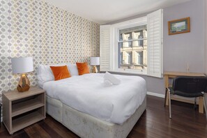 A neatly arranged bedroom with a bed, two bedside tables with lamps, a chair, a window with shutters, and a picture on the wall. - Flatzy - 3 Bedroom Apt in Heart of City Centre (Liverpool)