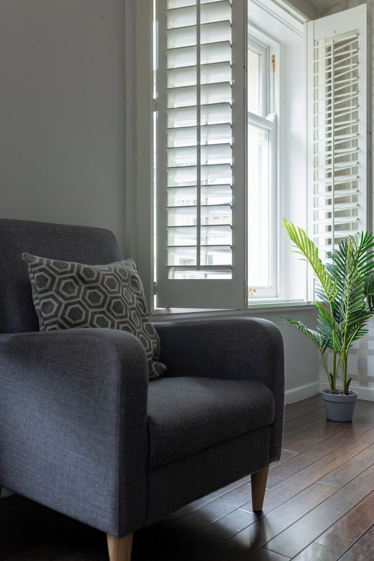 A grey armchair with a patterned pillow, positioned in front of a window with white shutters, and a potted plant to the right.