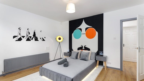 A modern bedroom with a large bed, a bedside table, a floor lamp, and a wall with a Beatles album cover. - Flatzy - Iconic Beatles and Liverpool Culture Home (Liverpool)