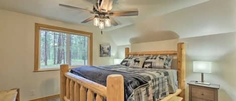 A bedroom with a wooden bed, a ceiling fan, a window with a forest view, and a nightstand with a lamp.
