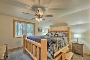 A bedroom with a wooden bed, a ceiling fan, a window with a forest view, and a nightstand with a lamp. - A-frame in the heart of Pinetop with AC (Pinetop-Lakeside)