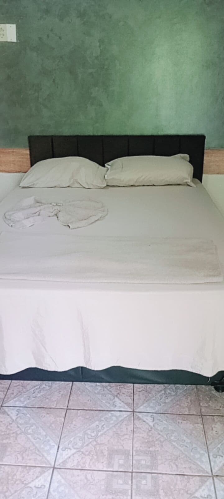 Una cama con una colcha blanca y almohadas, suelo de baldosas y pared verde.