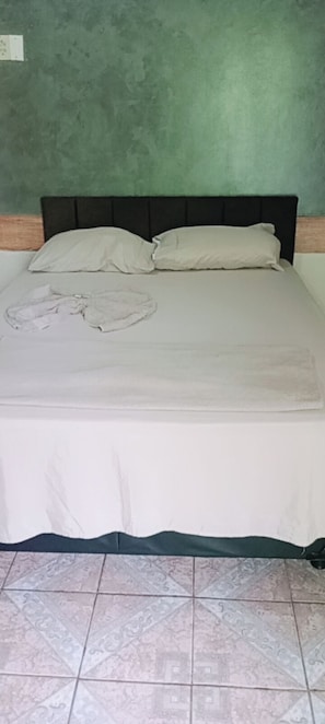 Uma cama com colcha e travesseiros brancos, chão de azulejos e parede verde.