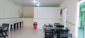 Uma área de refeições com mesas e cadeiras, um cesto de lixo e cozinha visível através de uma porta aberta.