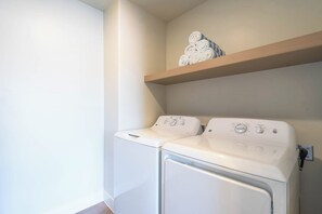 A laundry area with two white washing machines and a wooden shelf with rolled towels.
