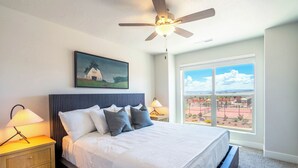 A bedroom with a bed, a ceiling fan, a painting, a nightstand with a lamp, and a window with a view.