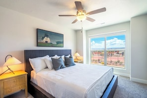 A bedroom with a bed, a ceiling fan, a painting, a nightstand with a lamp, and a window with a view.