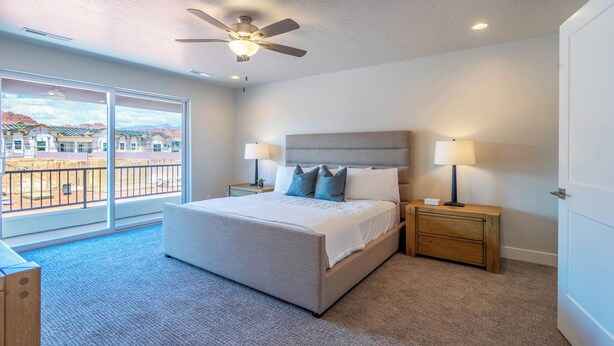 A bedroom with a bed, two bedside tables, a ceiling fan, and a view of a building through a large window.