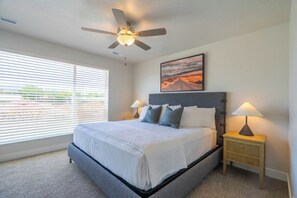 A bedroom with a bed, a ceiling fan, a nightstand with a lamp, and a window with blinds.