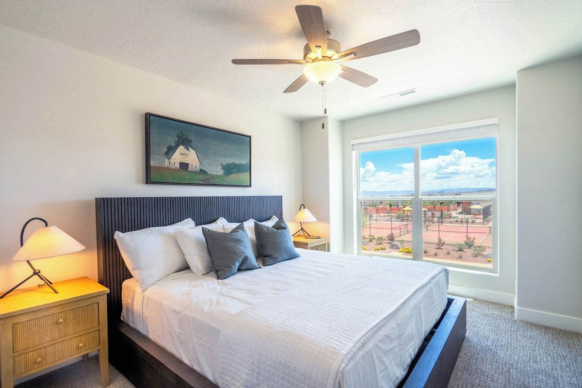 A bedroom with a bed, a ceiling fan, a nightstand with a lamp, a window with a view, and a painting on the wall.