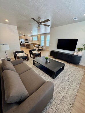 A modern living room with a sofa, coffee table, TV, and a dining area with chairs and a kitchen in the background.