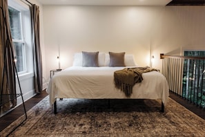 A bedroom with a large bed, a nightstand, a lamp, a window with curtains, and a balcony with a railing.