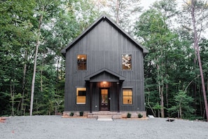 A two-story wooden house with a gabled roof, surrounded by a forest.
