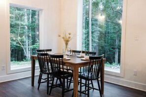 Un coin repas avec une table et des chaises en bois, de grandes fenêtres donnant sur la forêt et un vase rempli de fleurs séchées.