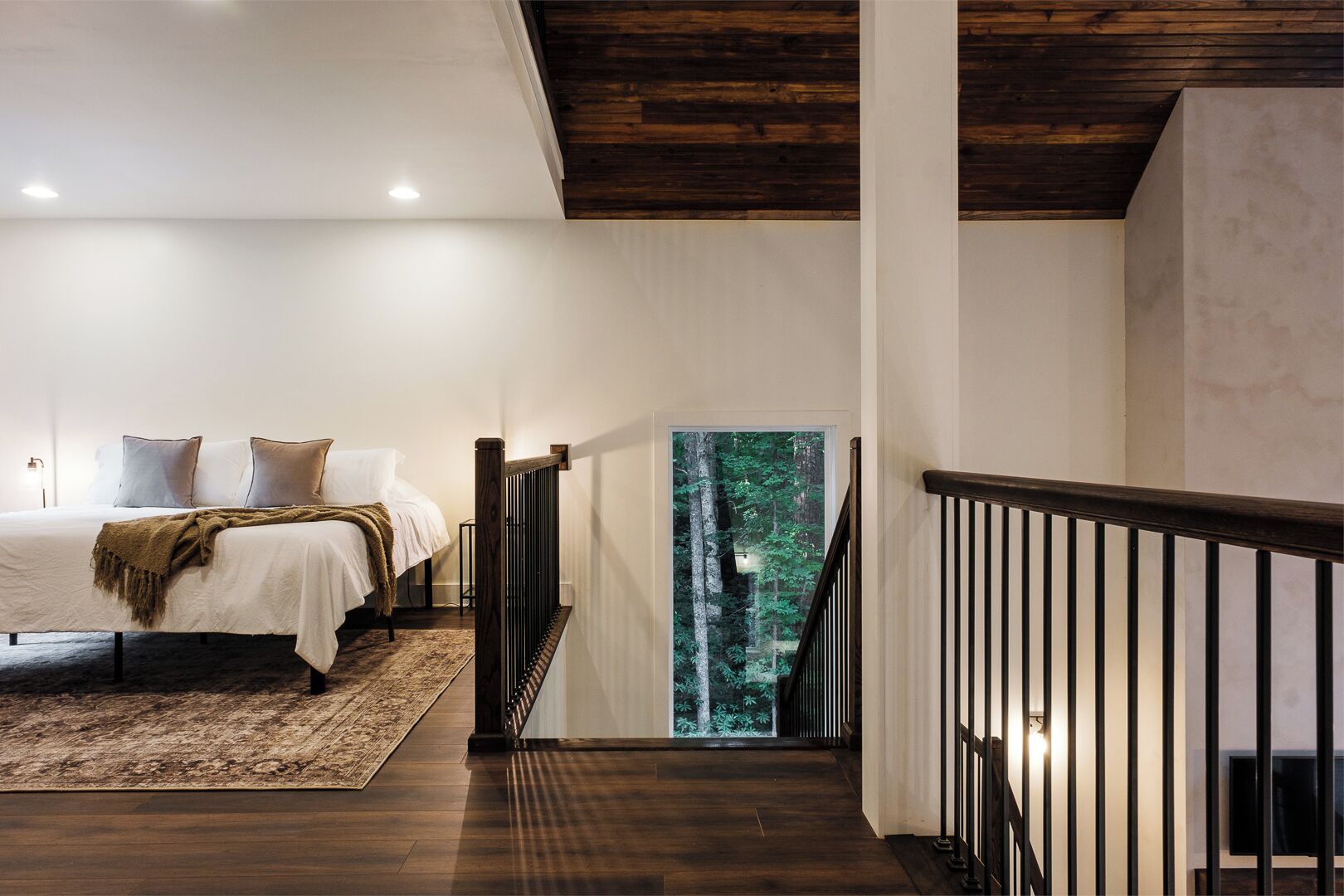 A bedroom with a large bed, a view of a forest through a glass door, and a wooden staircase.