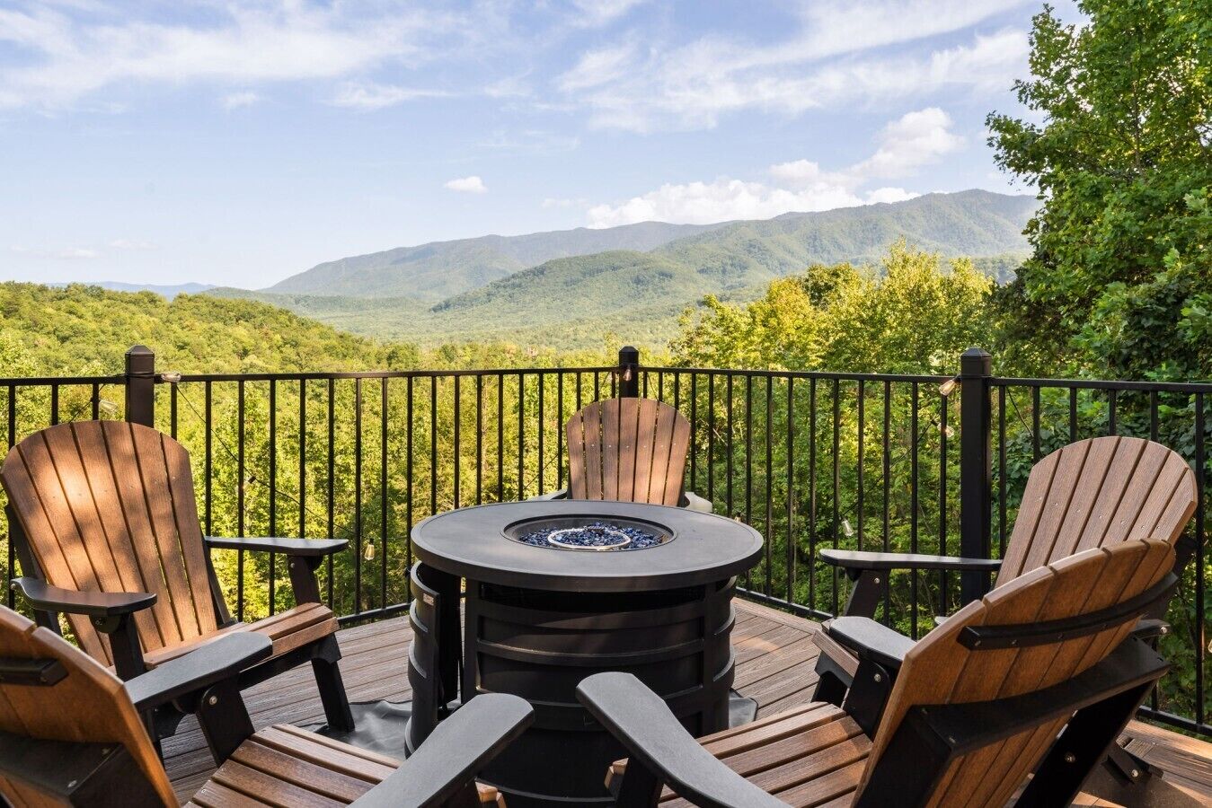 A deck with Adirondack chairs and a round table with a fire pit, overlooking a mountainous landscape.
